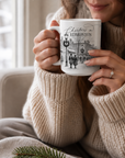 Woman in a cream knit sweater holding a Christmas in Edmonton mug with a festive town illustration beside a window.