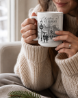 Woman in a cream knit sweater holding a Christmas in St. Albert mug with a festive town illustration beside a window.