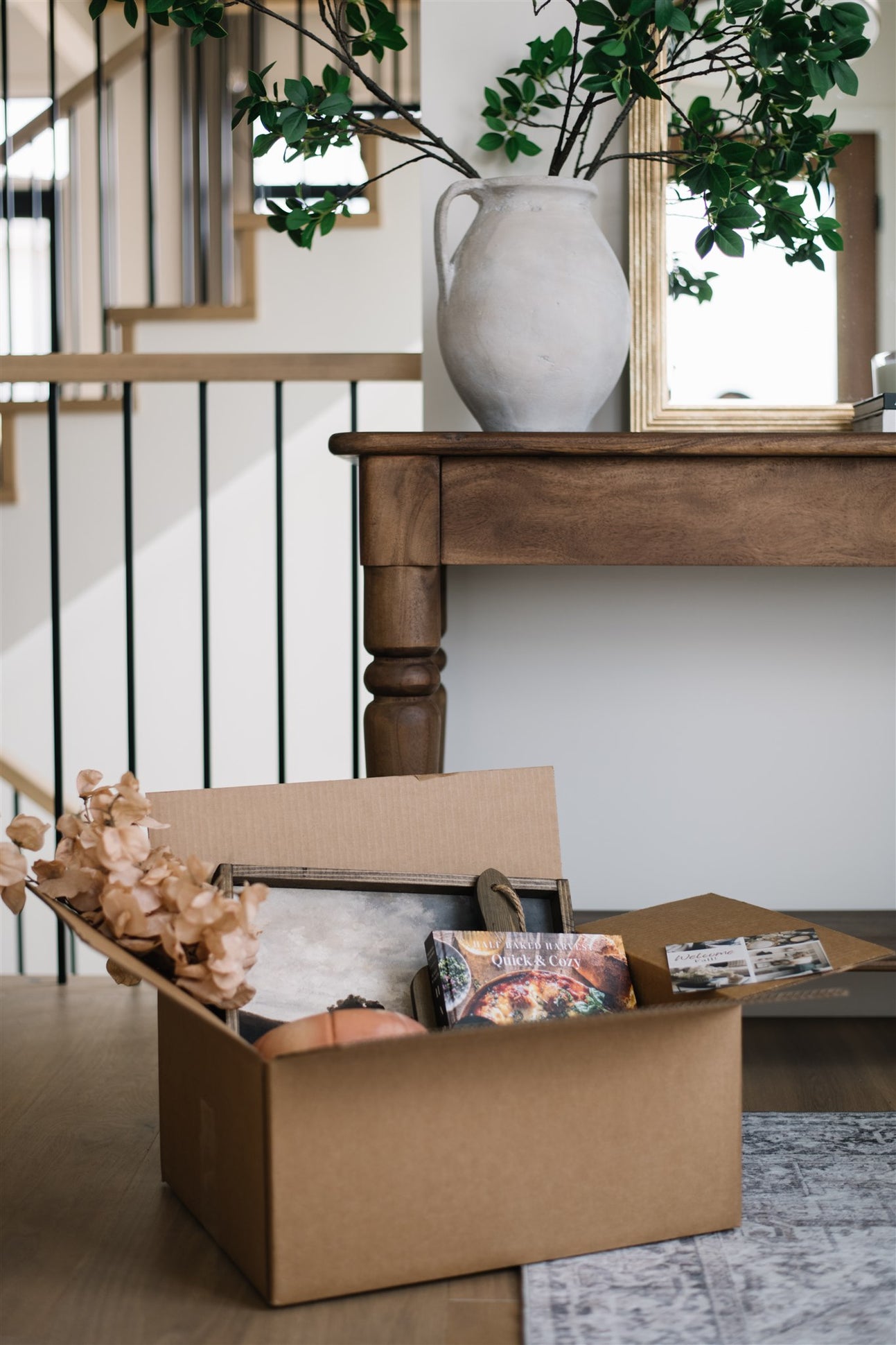 Open cardboard box with items on a wooden floor, with a vase and plant in the background.