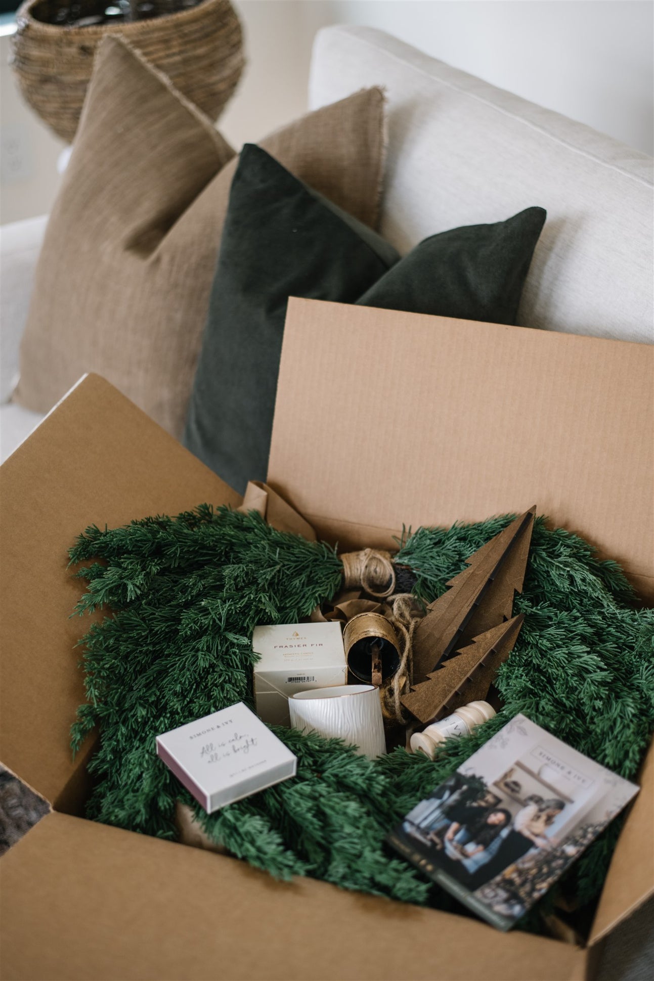 Box containing decorative items including a green wreath, candles, and a photo album on a couch with pillows.